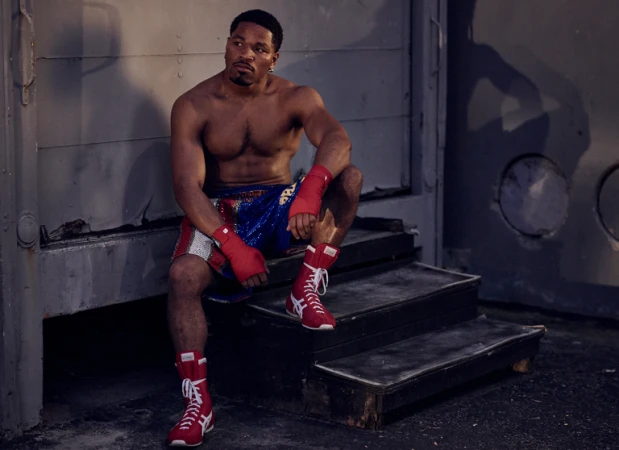 Muscular shirtless man in boxing shorts and red boots sits on gray steps, looking to the side, hands wrapped in red cloth.