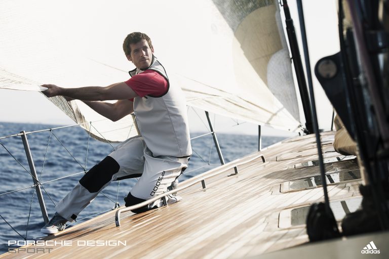 A man in activewear kneels on a sailboat, pulling a sail rope on a sunny day at sea, showcasing a blend of sport and style.