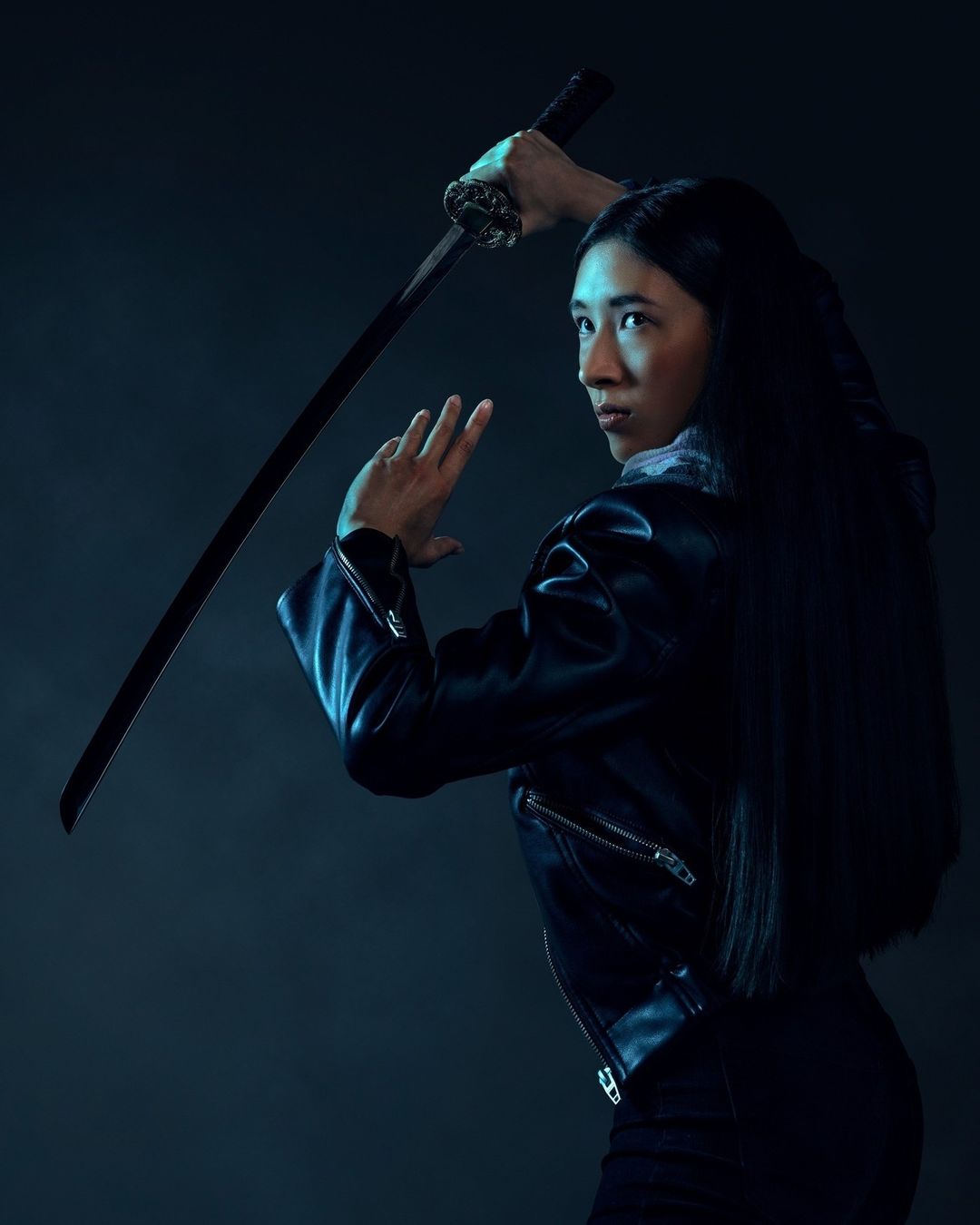 Young Asian woman in black leather jacket holding katana sword, looking over her shoulder in a dramatic studio shot.