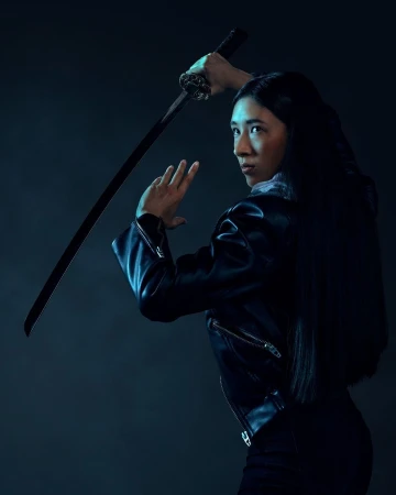 Young Asian woman in black leather jacket holding katana sword, looking over her shoulder in a dramatic studio shot.
