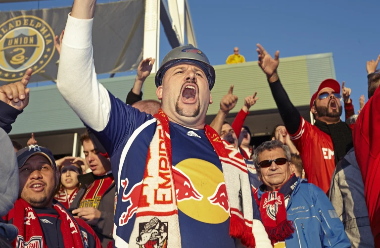 Enthusiastic sports fans cheer at an outdoor game, wearing team colors of red, white, and blue. A man in a helmet raises his fist.