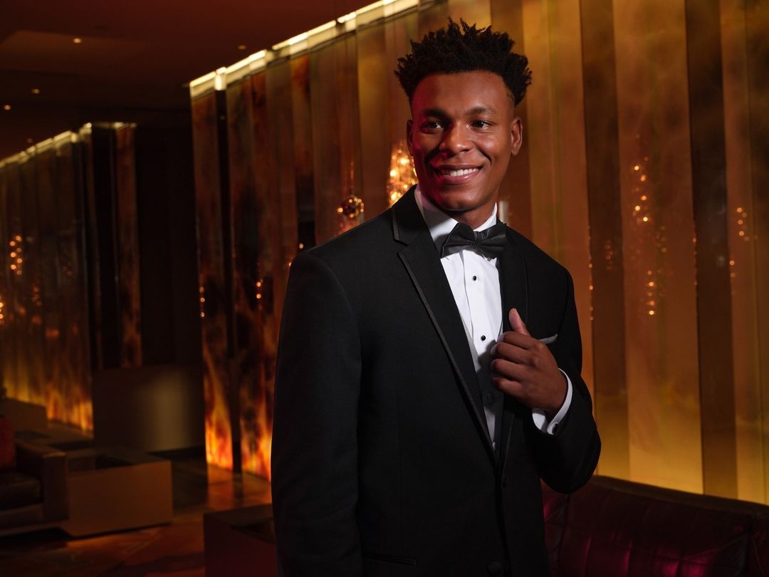 A smiling African-American man in a black tuxedo and bow tie poses in an upscale interior with a background of gold and brown tones.