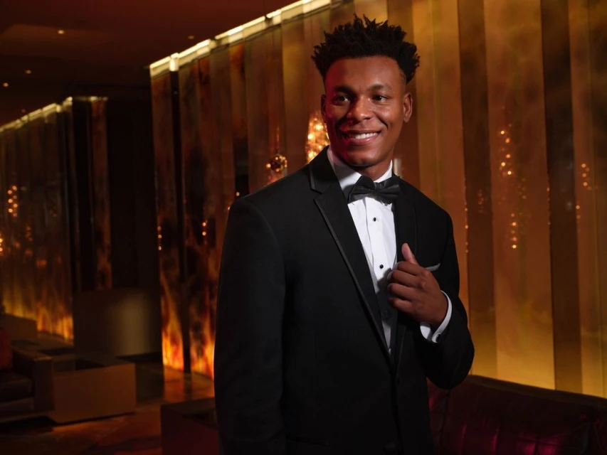 A smiling African-American man in a black tuxedo and bow tie poses in an upscale interior with a background of gold and brown tones.