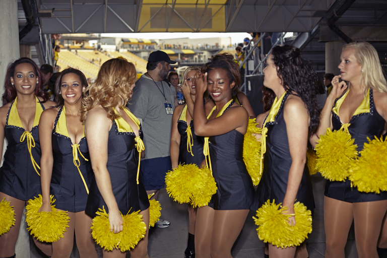Group of smiling cheerleaders in black and yellow uniforms holding yellow pompoms in a stadium hallway, with stadium seating visible.