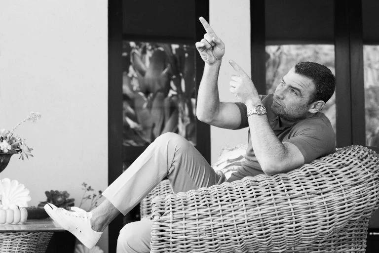 Man sitting in a wicker chair points upwards; black and white photo with relaxed pose, wearing casual clothes and a watch, indoor setting.