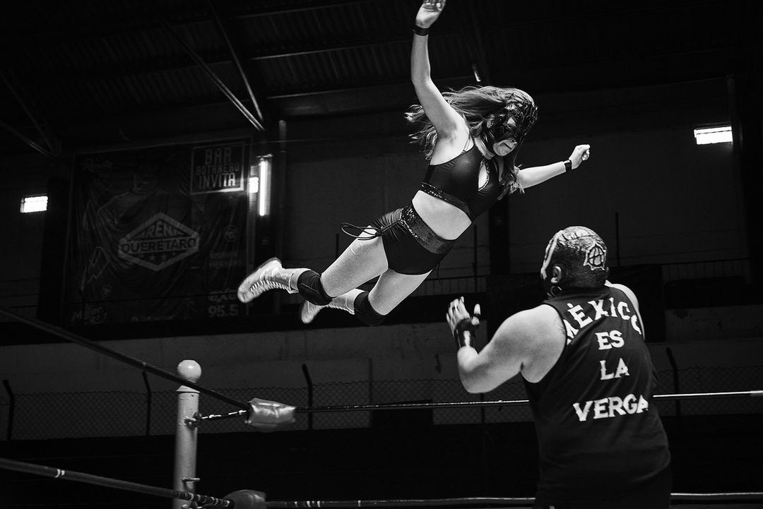 A black and white shot of a female wrestler in mid-air above a ring, with another wrestler below her, in an indoor stadium.