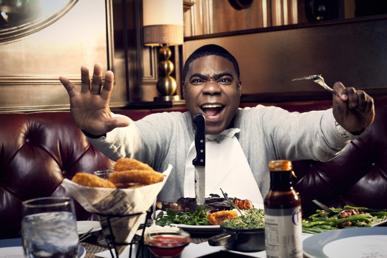 Man at a table covered with a large meal, he is smiling with arms raised, holding a fork. He is dressed in grey with a white bib.