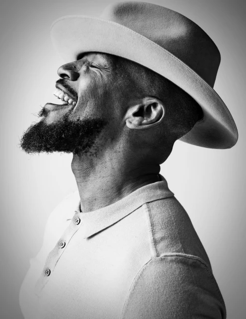 Black and white portrait of a Black adult male wearing a fedora and polo shirt with a joyful expression, head tilted back.