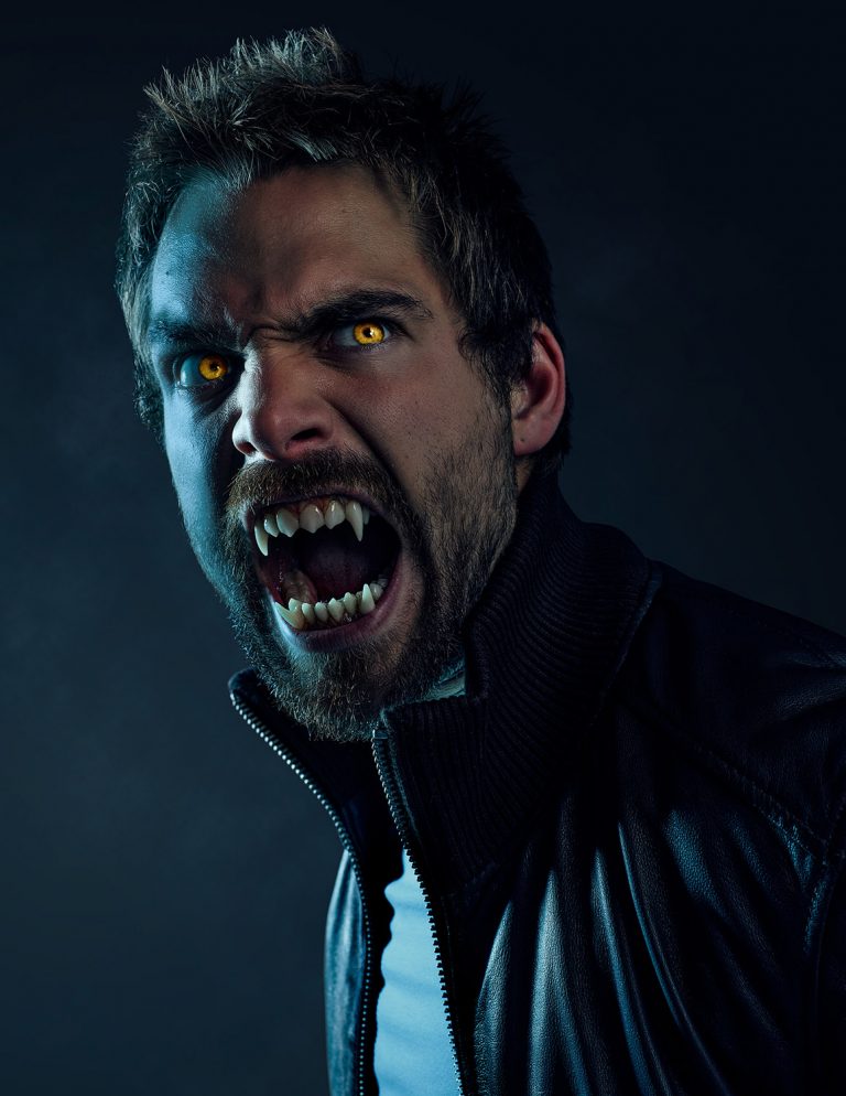 Close-up of a male actor with golden eyes, long fangs, bared teeth, and a dark leather jacket against a dark, smoky background.