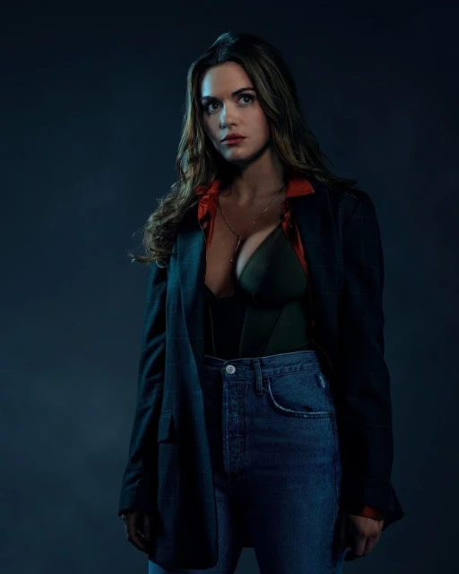 Woman with long brown hair poses indoors wearing a black blazer, blue jeans, and a dark top, against a dark blue backdrop.