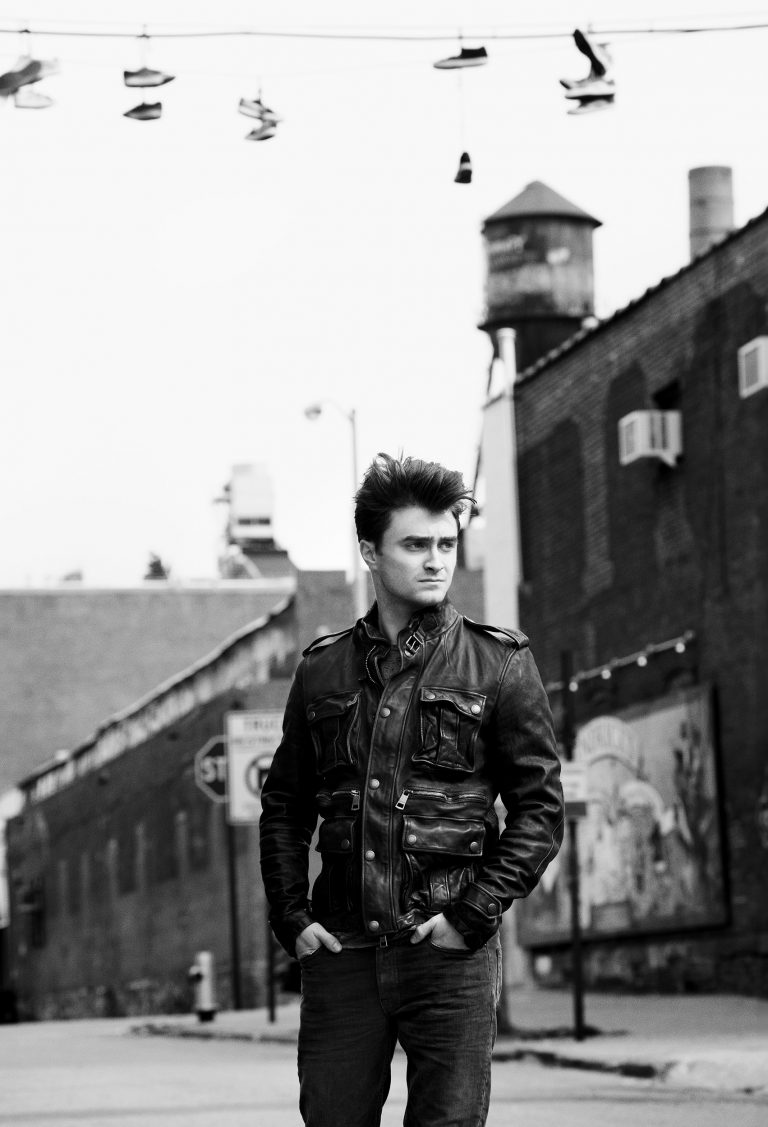 Monochrome portrait of a man in an urban setting, wearing a leather jacket, with shoes hanging overhead, creating a dramatic editorial style.