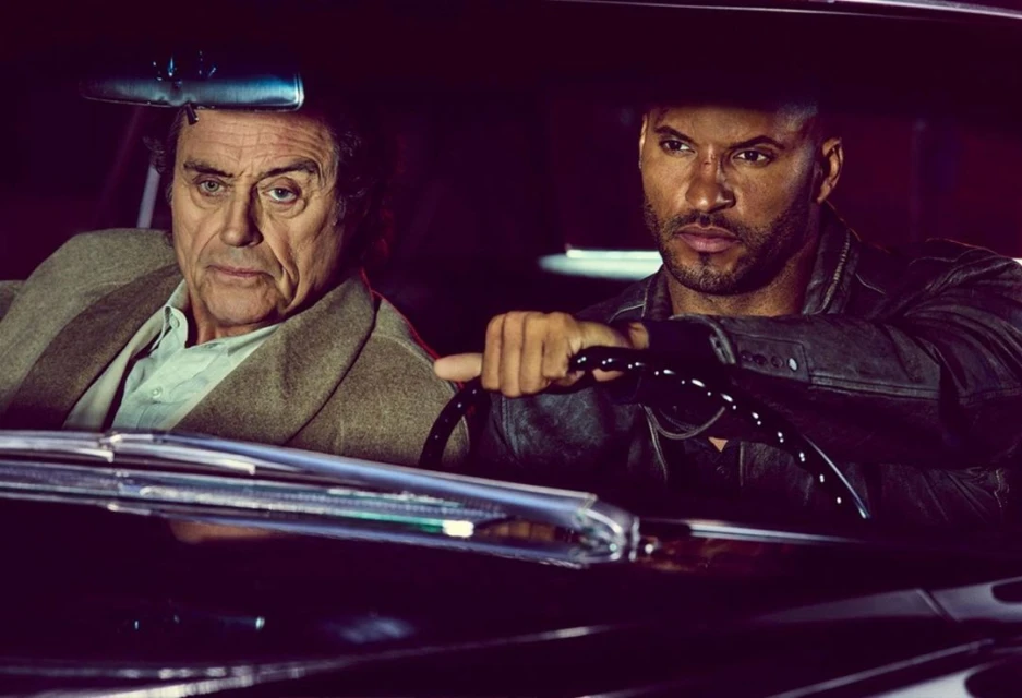 An older Caucasian man and a younger Black man are seated in a vintage car with dark leather seats, under dim lighting.