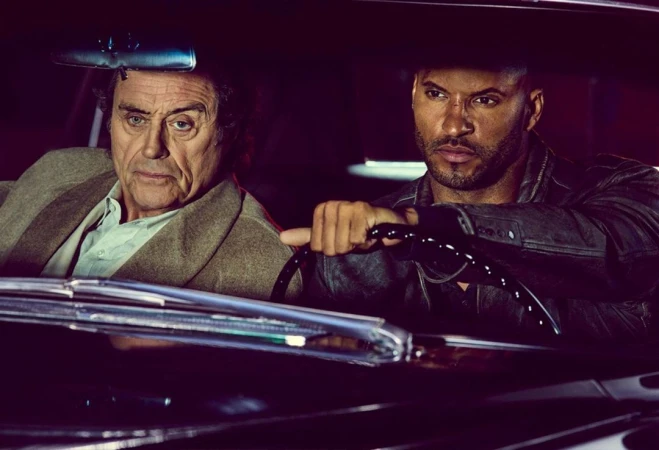 An older Caucasian man and a younger Black man are seated in a vintage car with dark leather seats, under dim lighting.