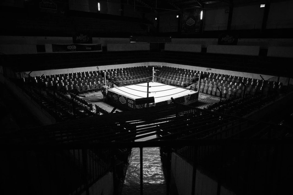 Black and white photograph of a boxing ring in an empty arena with rows of seating, low light, and shadows.