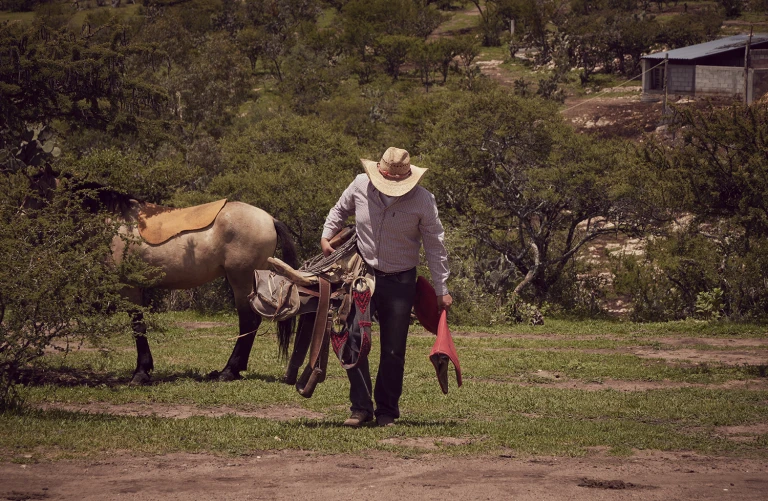A cowboy in a hat carries a saddle and a red cloth in a field, with a horse partially visible, in a rural setting with trees and a building.