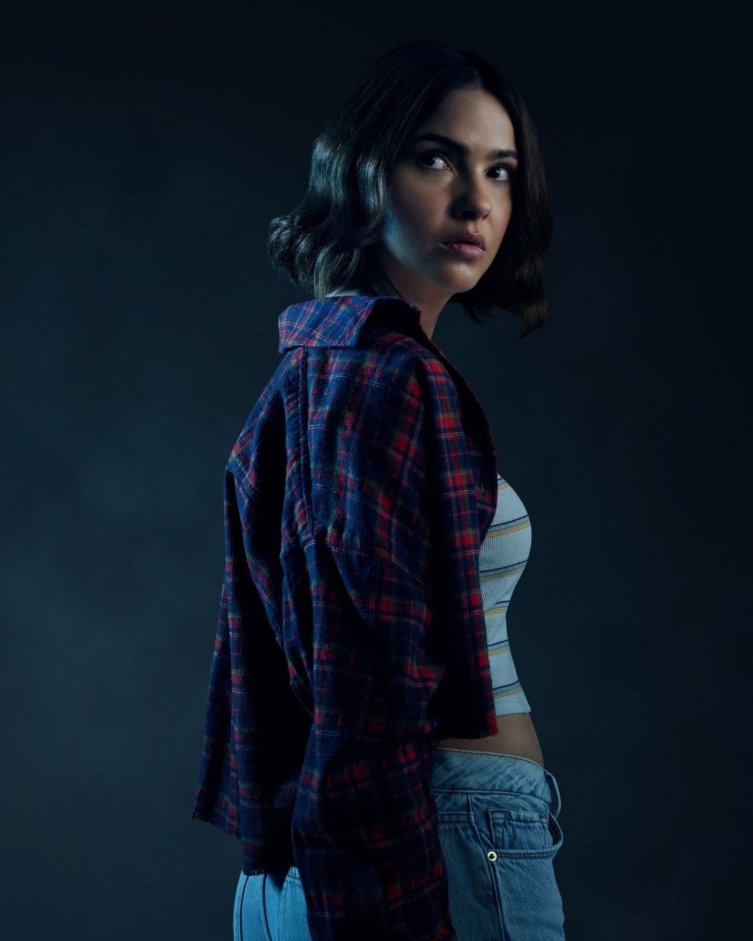 Portrait of a young woman with dark hair looking over her shoulder, wearing a striped top and plaid shirt against a dark background.