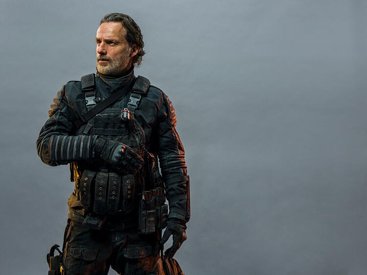 Man with beard in tactical gear, wearing a black uniform with a vest and pouches, standing in front of a plain grey background.