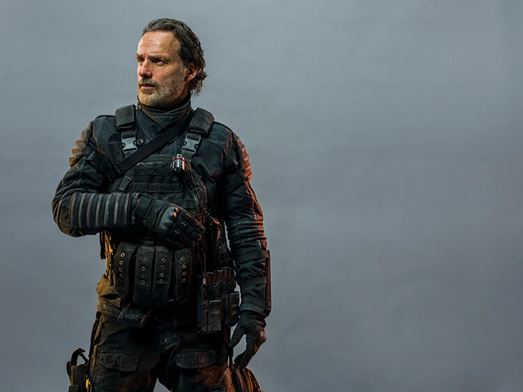 Man with beard in tactical gear, wearing a black uniform with a vest and pouches, standing in front of a plain grey background.