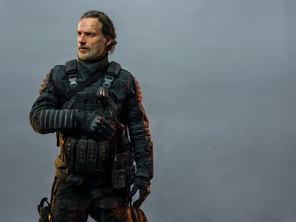 Man with beard in tactical gear, wearing a black uniform with a vest and pouches, standing in front of a plain grey background.