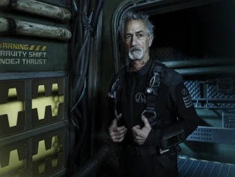 Older man with grey hair and beard in a black jumpsuit and harness standing inside an industrial spaceship interior
