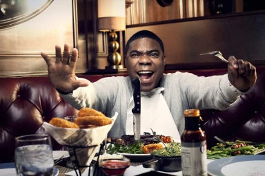 Tracy Morgan sitting at a red leather booth in a restaurant holding up a fork and knife while smiling broadly