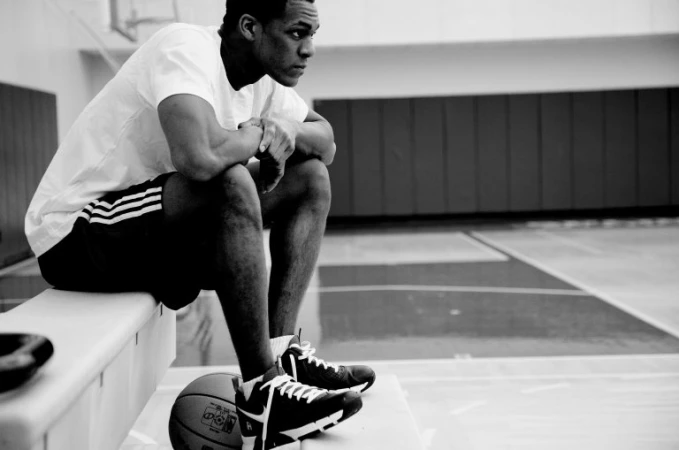 Black and white portrait of a young man seated on a bench resting arms on knees in an indoor sports venue