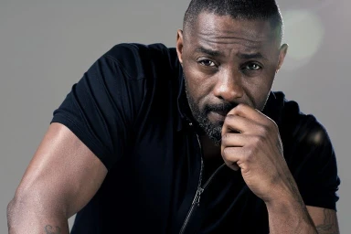 Portrait of Idris Elba wearing a black zipped top with hand near mouth against a neutral grey studio background