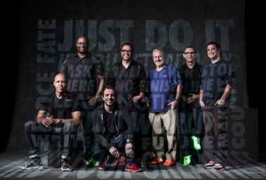 Group portrait of six men posing against a dark textured wall with faint typography by James Dimmock.