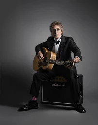 Portrait of Roger Daltrey in a black tuxedo playing an acoustic guitar while seated on a Marshall amplifier