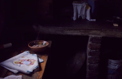 A rustic wooden table in a dimly lit room holds a ceramic bowl of beans and a folded white cloth decorated with an embroidered image.