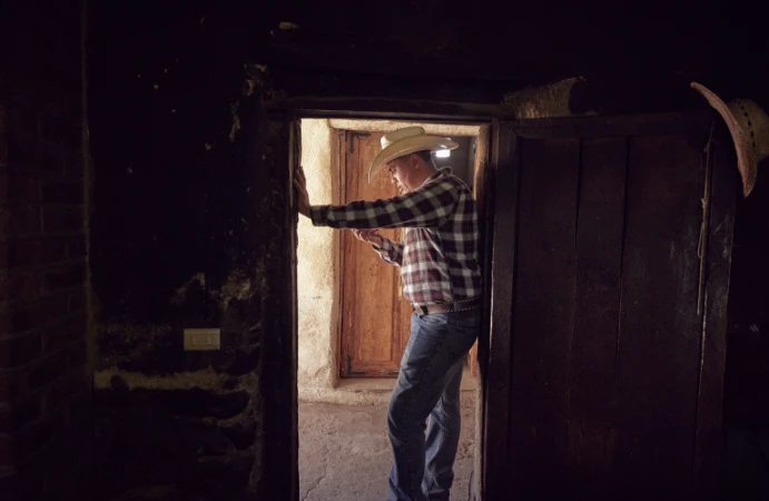 Man wearing a cowboy hat and plaid shirt standing in a rustic doorway holding a phone