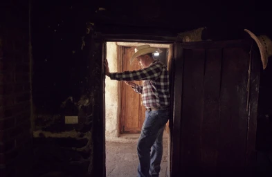 Man wearing a cowboy hat and plaid shirt standing in a rustic doorway holding a phone