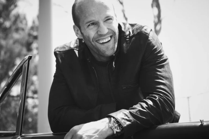 Portrait of Jason Statham smiling while leaning against a convertible car, wearing a dark leather jacket.