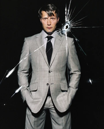 Mads Mikkelsen wearing a grey plaid suit and dark tie against a black background with a shattered glass effect over his shoulder.