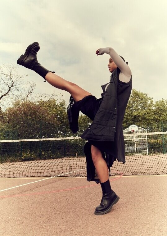A person poses with their leg in the air, wearing a black vest, skirt, socks, and boots, on a sports court with a net behind them.