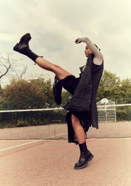 A person poses with their leg in the air, wearing a black vest, skirt, socks, and boots, on a sports court with a net behind them.