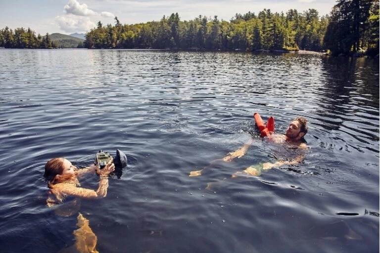 Woman photographs man swimming in a lake surrounded by trees and mountains on a sunny day with clear skies and blue water.