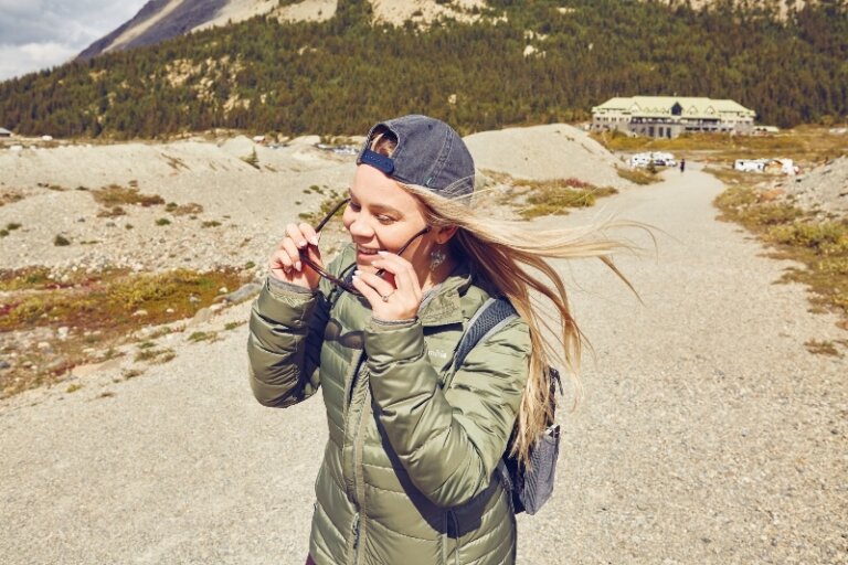Woman in green puffer jacket holding sunglasses, stands on path in mountainous landscape. Mountain lodge is visible in background.