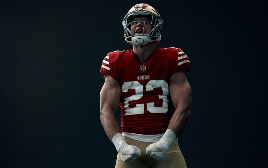 An American football player in a red uniform, helmet, and white hand wraps clenches his fists with a determined facial expression.