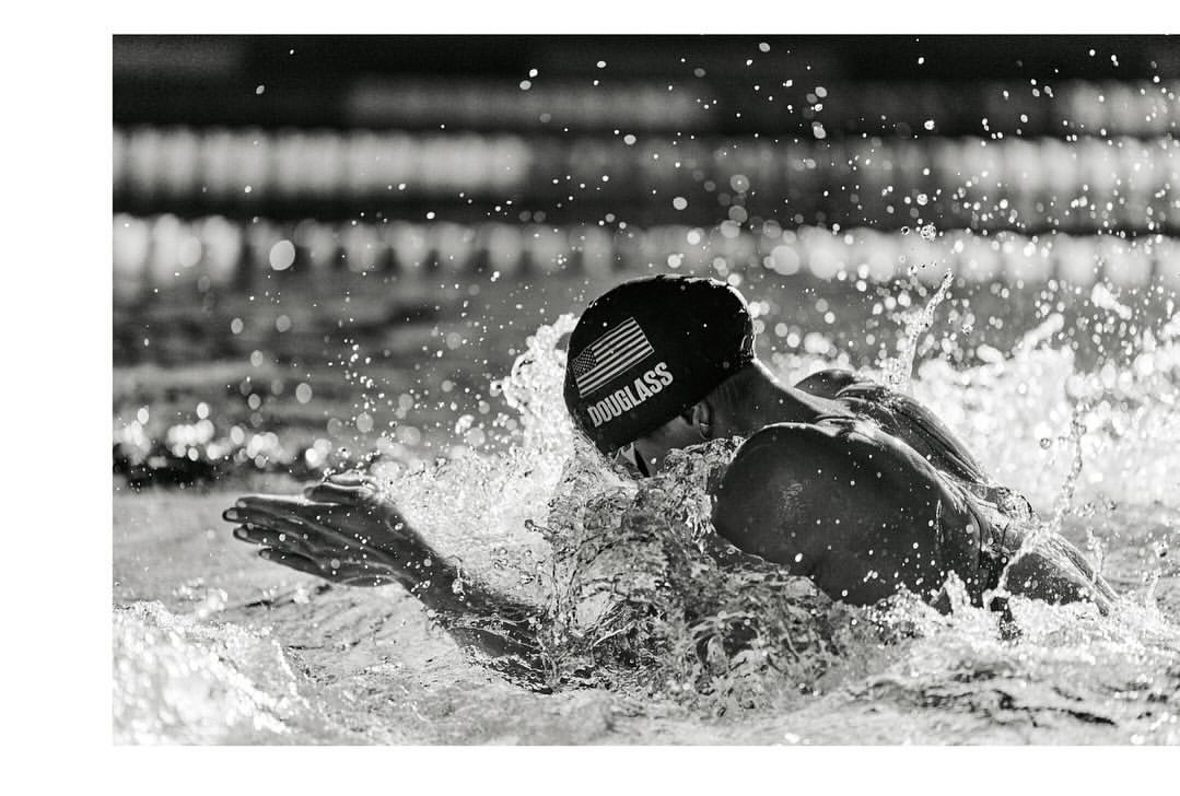 Monochromatic action shot of a swimmer with swim cap and goggles in water, creating splash, with tile background and USA flag.