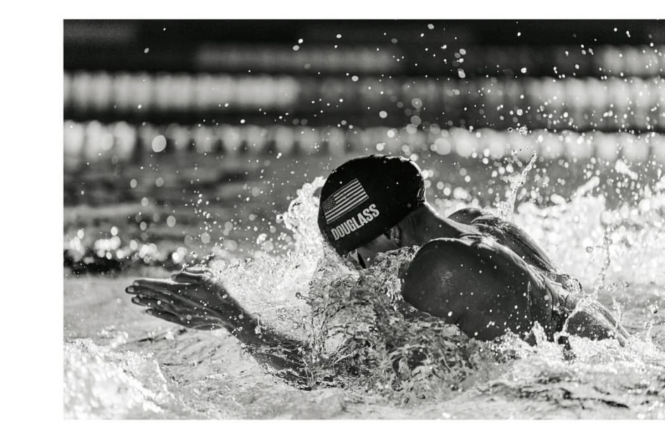Monochromatic action shot of a swimmer with swim cap and goggles in water, creating splash, with tile background and USA flag.