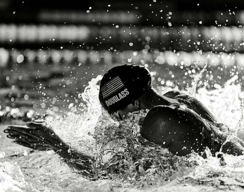 A swimmer wearing a cap with an American flag is captured mid-stroke in a black-and-white photo, surrounded by splashing water.