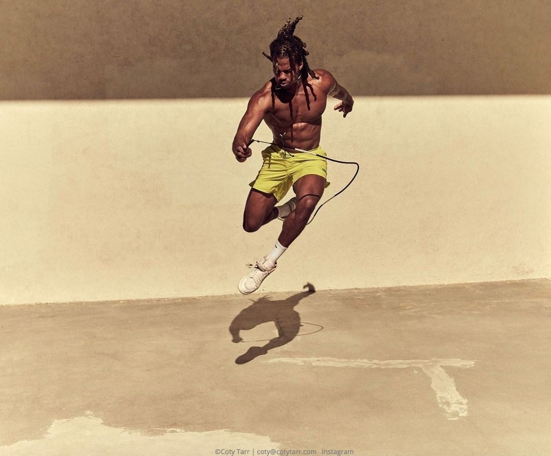 Athletic man with dreadlocks is caught mid-air jump roping in bright yellow shorts, wearing white sneakers, outdoors on a sunny day.