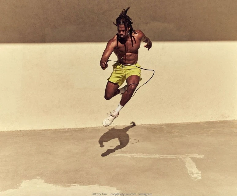 Athletic man with dreadlocks is caught mid-air jump roping in bright yellow shorts, wearing white sneakers, outdoors on a sunny day.