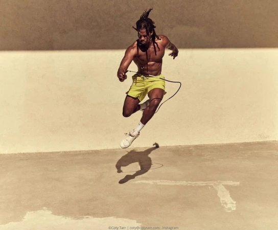 Athletic man with dreadlocks is caught mid-air jump roping in bright yellow shorts, wearing white sneakers, outdoors on a sunny day.