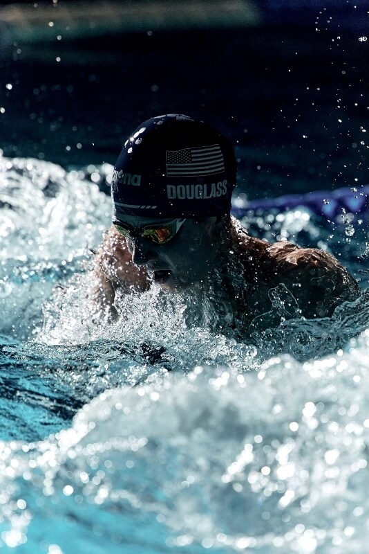 Man swimming butterfly stroke with American flag swim cap and goggles in a pool with splashing water, dark background.