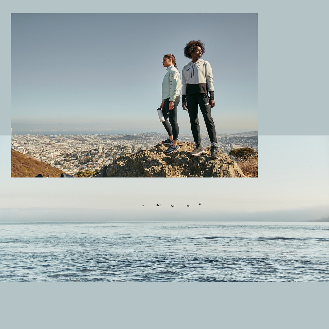 Two women in athletic wear stand on a rocky hilltop overlooking a city skyline and the ocean under a clear sky, enjoying an outdoor scene.