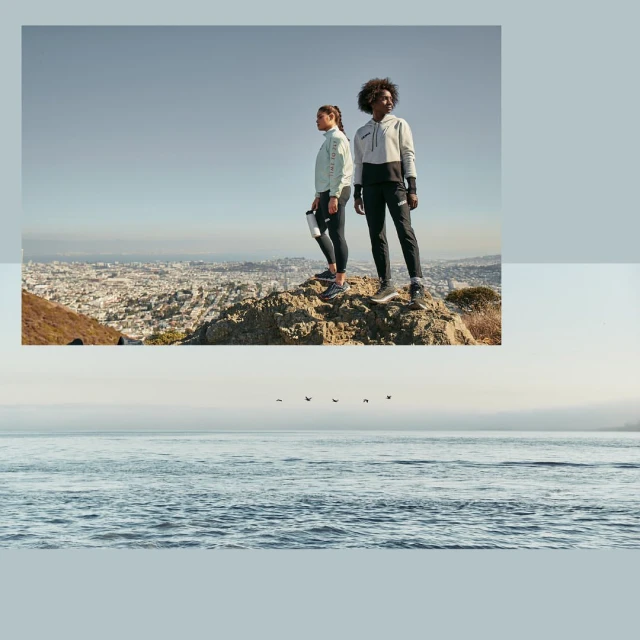 Two women in athletic wear stand on a rocky hilltop overlooking a city skyline and the ocean under a clear sky, enjoying an outdoor scene.