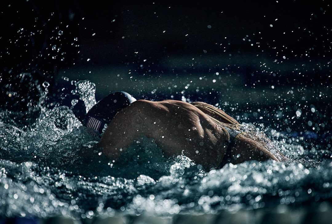 Focused adult male swimmer in cap and goggles propelling through splashing water, lit dramatically against a dark background in a pool.