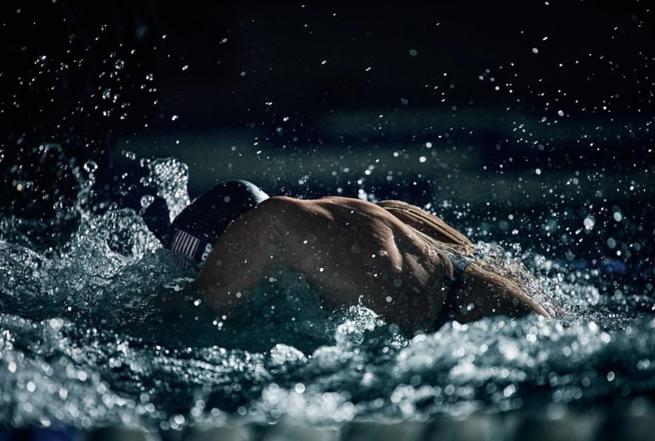 Focused adult male swimmer in cap and goggles propelling through splashing water, lit dramatically against a dark background in a pool.